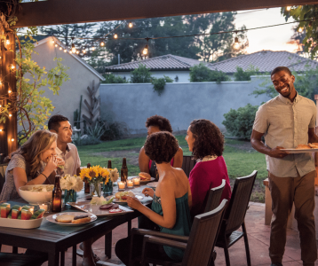 A group of six people share an outdoor dinner under string lights, seated around a table with food, drinks, candles, and flowers in a garden setting.