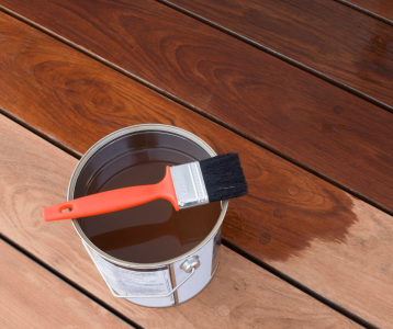 A paintbrush rests on the rim of a can of wood stain beside a wooden deck surface showing a clear contrast between the unstained and freshly stained sections.