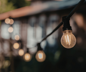 A close‑up of outdoor string lights with one bulb in sharp focus and several blurred bulbs in the background against greenery and a softly lit building.