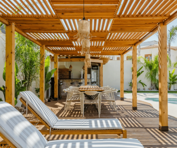 A spacious outdoor living area with a wooden pergola, cushioned lounge chairs, a long dining table, woven pendant lights, and an outdoor kitchen beside a pool.