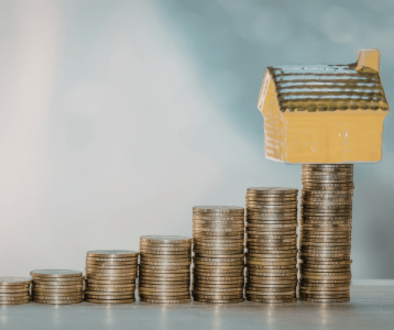 Stacks of coins arranged in ascending height with a small yellow ceramic house placed on the tallest stack, symbolizing increasing property value.