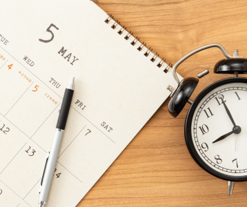 A desk setup with a spiral‑bound calendar open to May, a pen resting on the page, and a black analog clock.